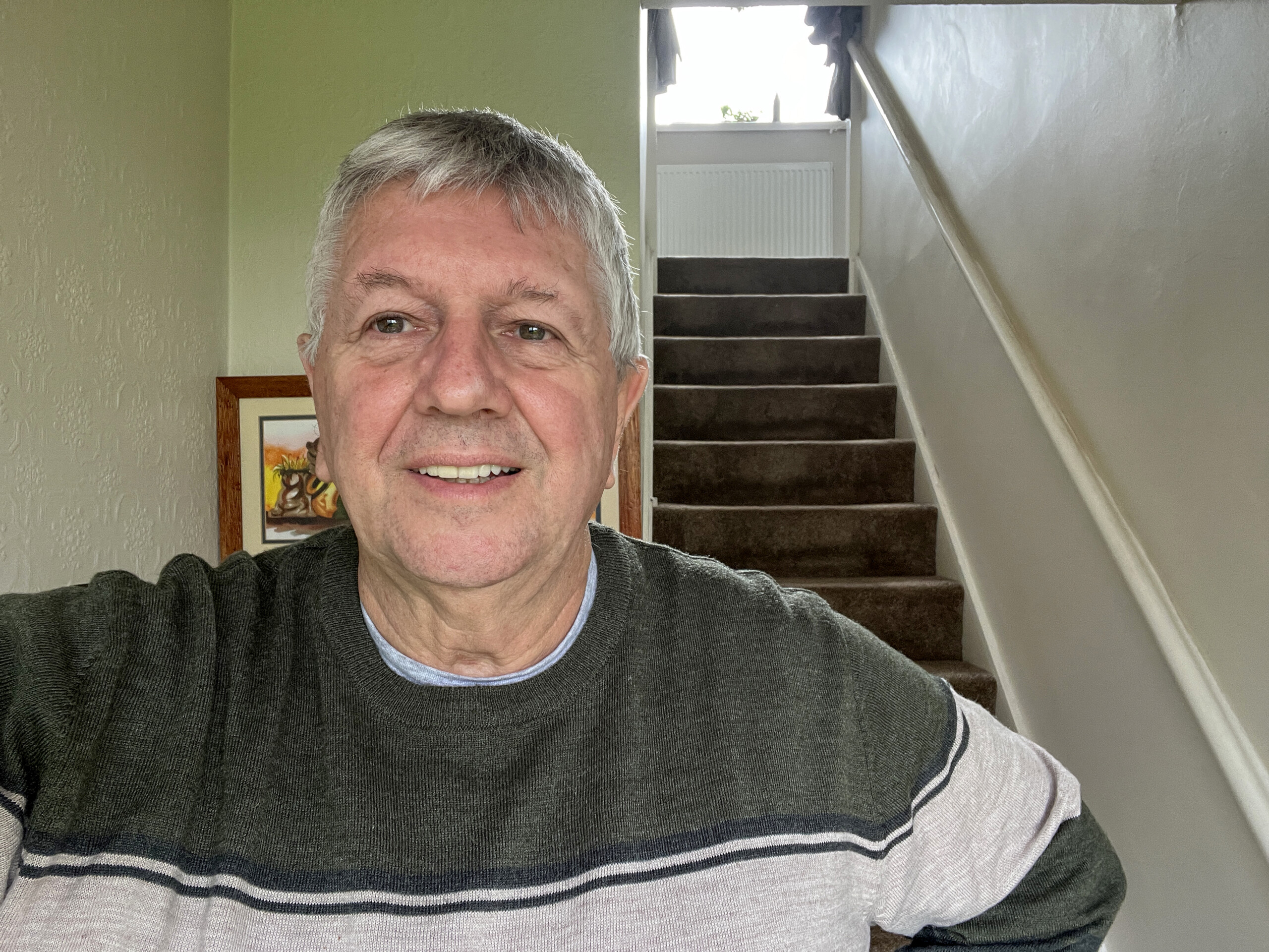 An older man with grey hair and a stripey jumper takes a smiling selfie in his home. Behind him you can see his hallway and stairs.