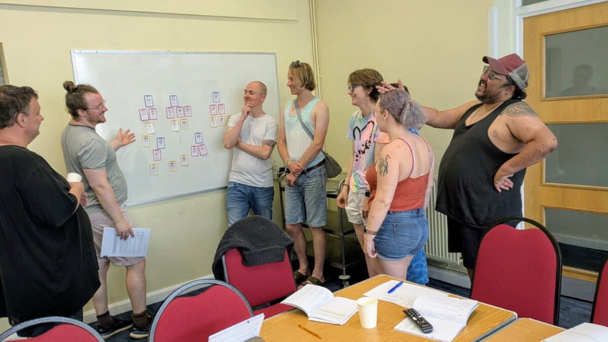 A money coachig group of people smiling, laughing and pointing at a whiteboard with sorted boxes on it.