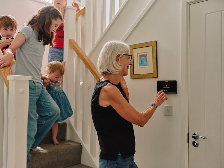 An older woman adjusts the settings of the heatpump while behind her her grandchildren descend the stairs.