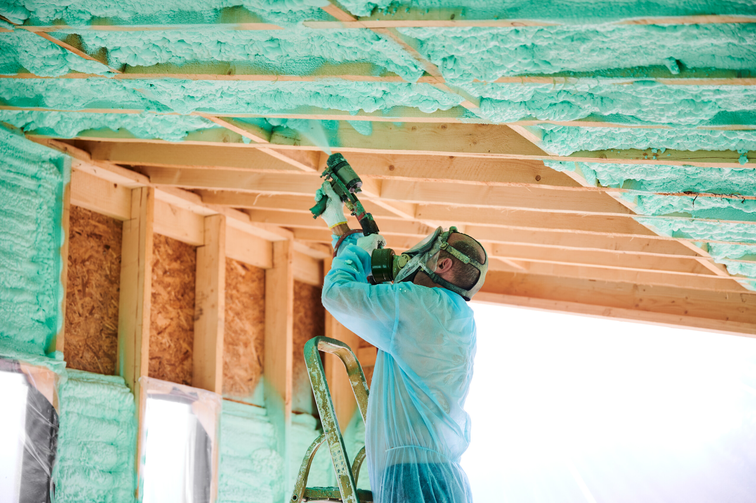 A man in protective clothing, face mask and breathing apparatus, sprays insulation foam on to the inside of a roof.