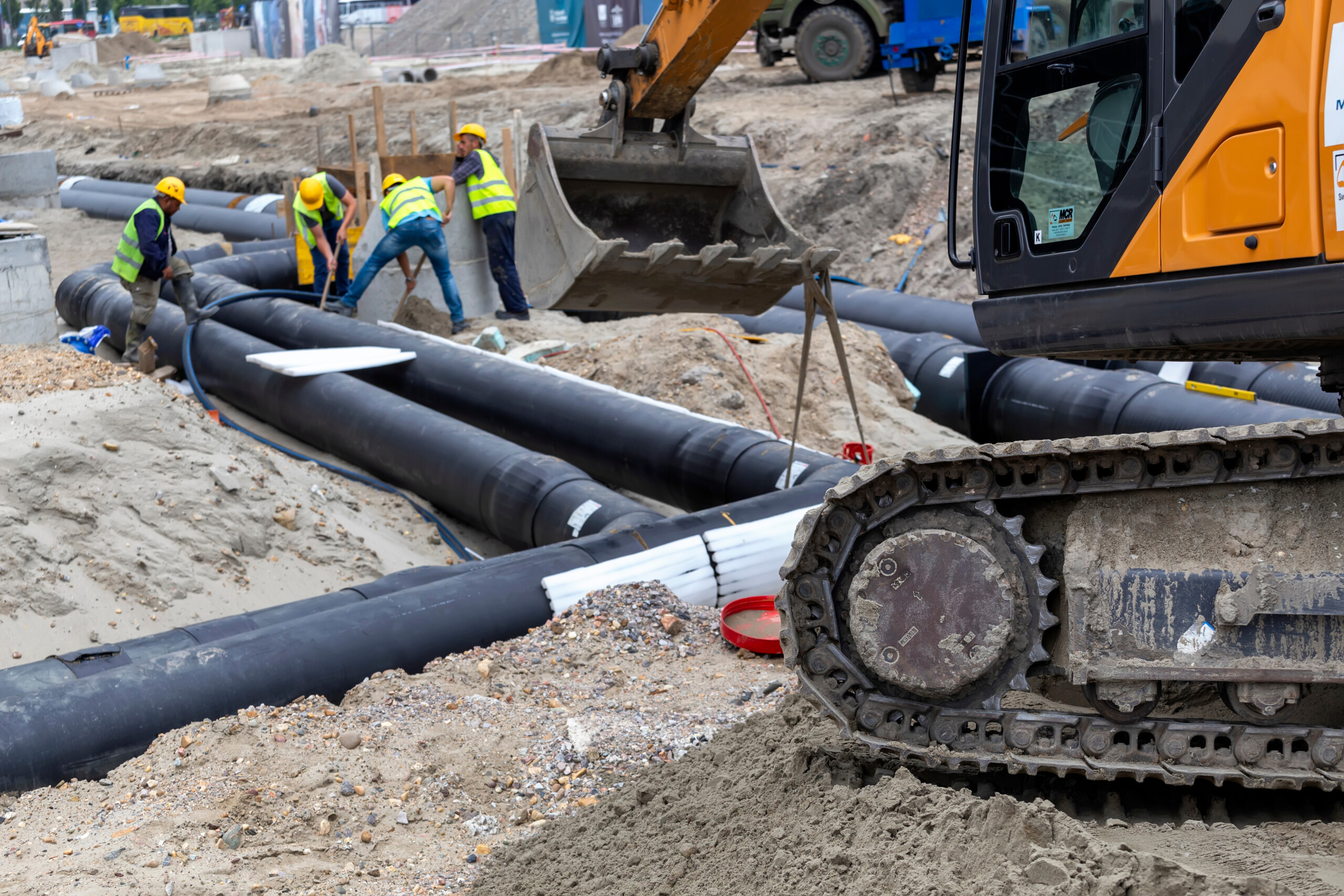 An excavator in the foreground with workers laying down heat network pipes in the background