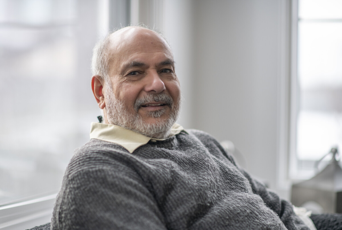 A beautiful smile from an elderly senior man. He is wearing a grey sweater and is seated in front of big bright windows at his home.