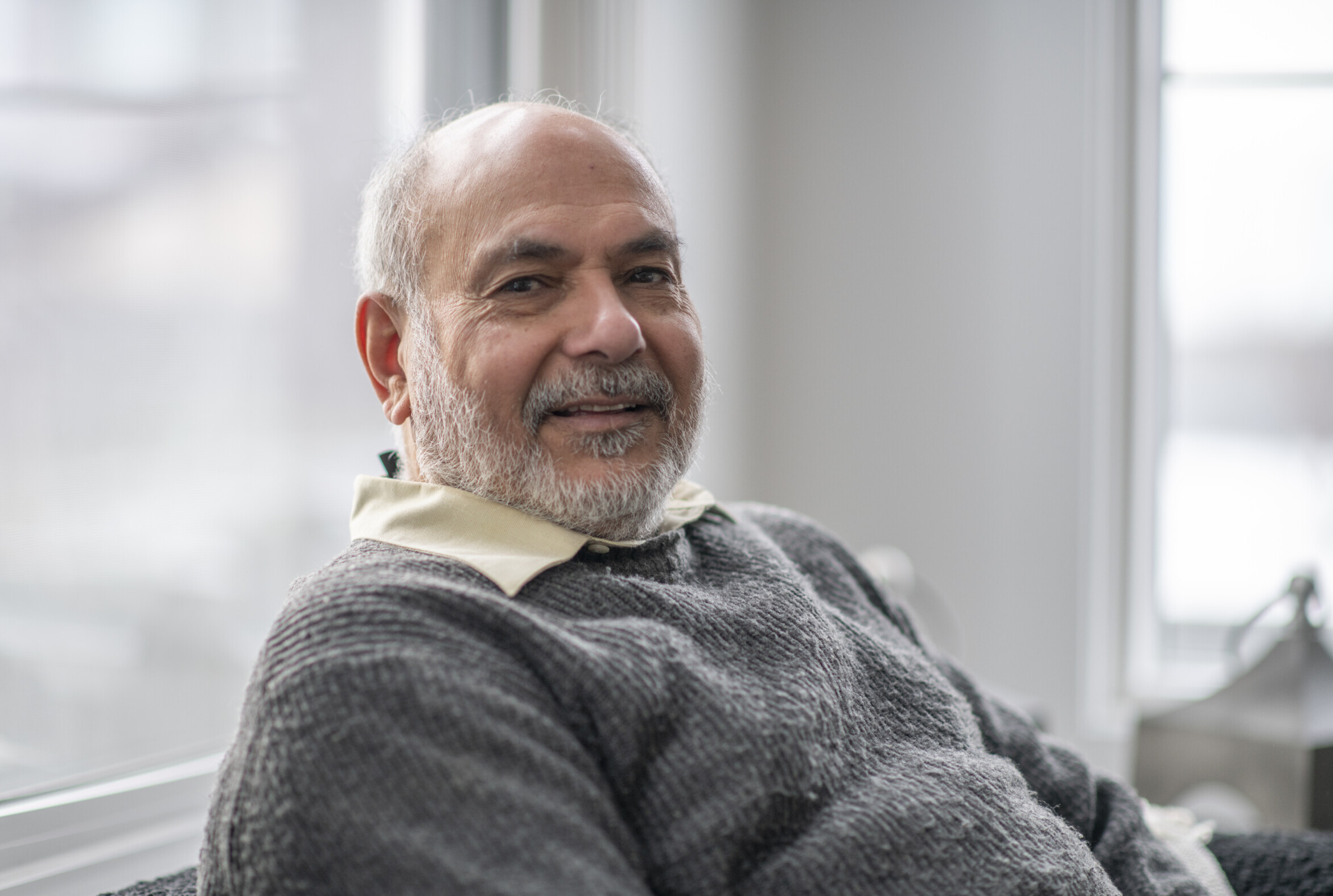 A beautiful smile from an elderly senior man. He is wearing a grey sweater and is seated in front of big bright windows at his home.