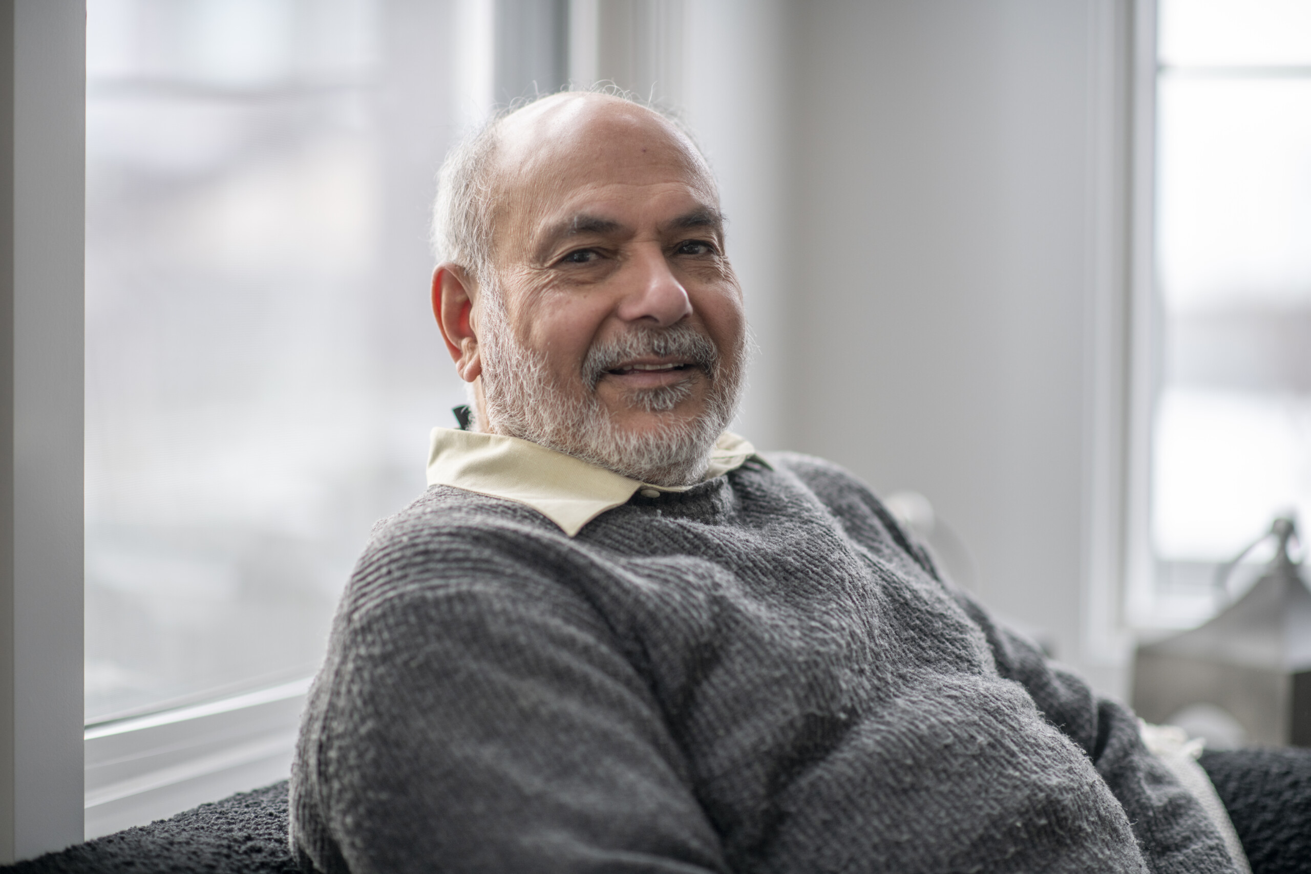 A beautiful smile from an elderly senior man. He is wearing a grey sweater and is seated in front of big bright windows at his home.