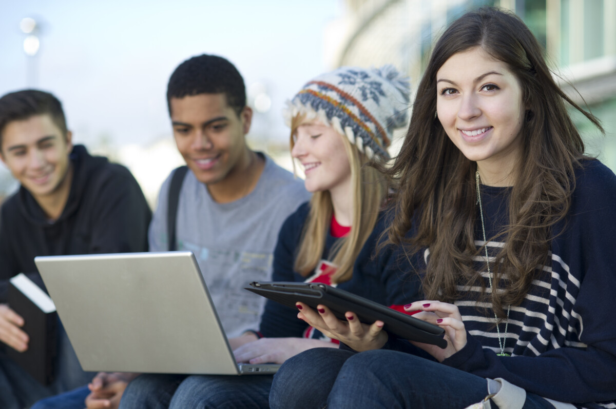Four young people sitting in a row and smiling, some looking at laptops and tablets.