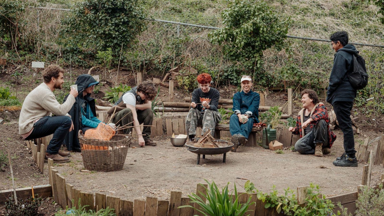 A group of young people sit around a firepit out in nature.