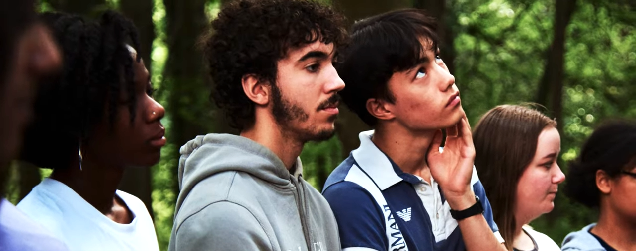 A row of young people stand outside in nature. They're all looking out into the distance, looking thoughtful.
